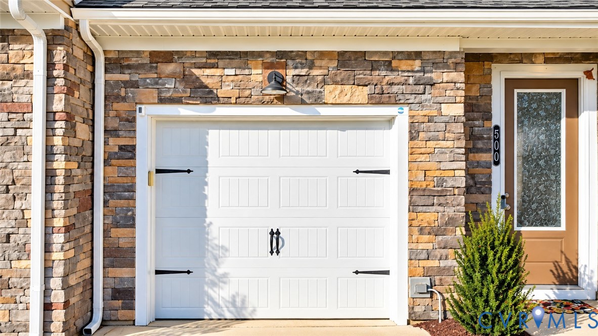 Multi-textured stone facade featuring a white carriage-style garage door with decorative black hardware, an exterior wall-mounted gooseneck light fixture, and a glass-paneled entry door Multi-textured stone facade featuring a white carriage-style garage door with decorative black hardware, an exterior wall-mounted gooseneck light fixture, and a glass-paneled entry door