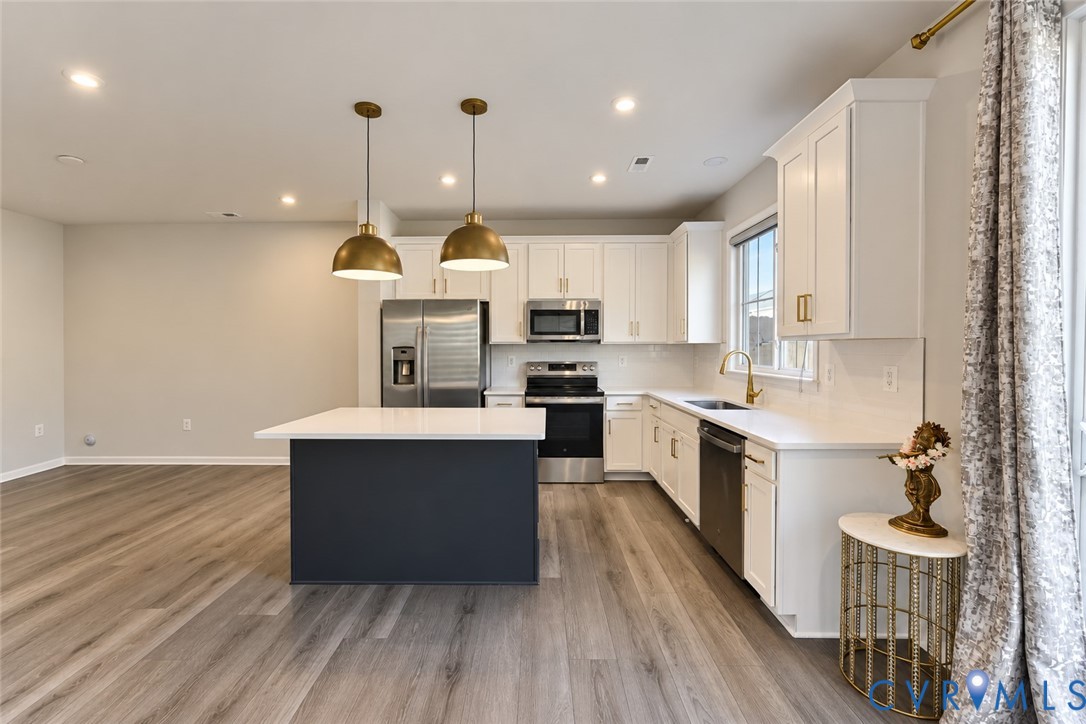 Kitchen featuring white cabinetry, wood-finish flooring, a central island with a solid-surface countertop, stainless steel appliances, and brass-tone pendant lighting