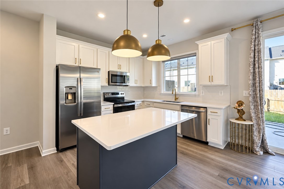 Kitchen featuring white cabinetry, an island with a quartz countertop, stainless steel appliances, dual brass-finish pendant lights, and wood-finish flooring