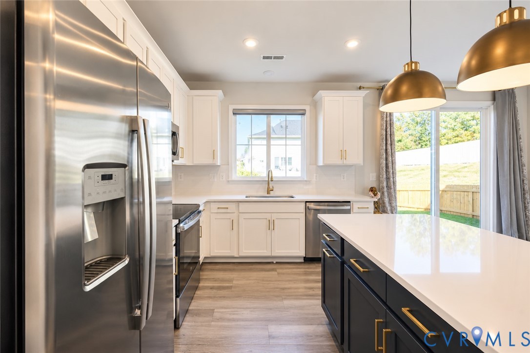 Kitchen featuring white upper cabinetry, a dark-toned center island with a quartz countertop, stainless steel appliances, brass-finish pendant lighting, and wood-finish flooring