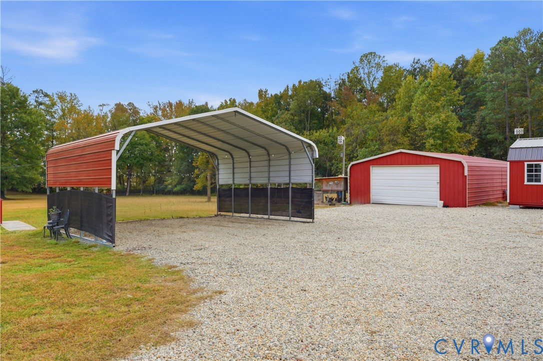Metal-roofed carport featuring a gravel-surfaced drive, red steel-sided outbuilding with a roll-up garage door, and a wooded perimeter