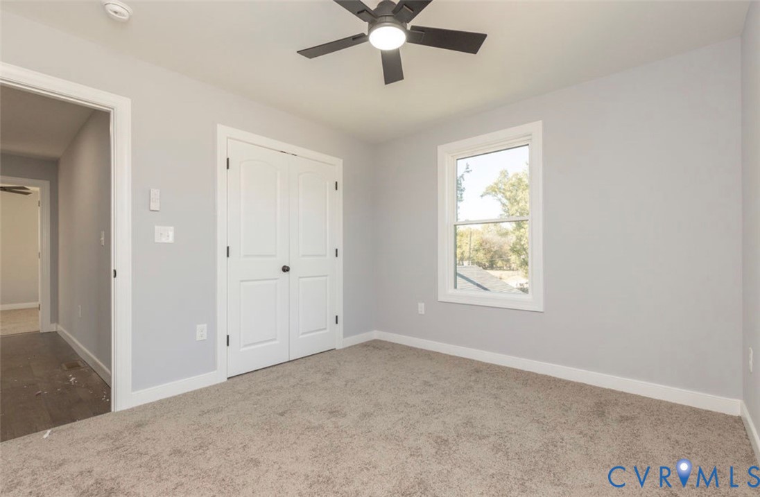 Neutral-toned interior space featuring neutral wall-to-wall carpeting, double-panel closet doors, a multi-pane window, and a ceiling-mounted light fixture with an integrated fan