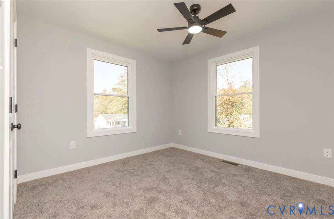 Neutral-toned interior featuring wall-to-wall carpeting, dual double-hung windows, and a matte black ceiling fan with integrated lighting