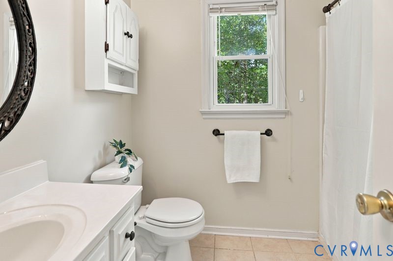 Bathroom featuring a white vanity cabinet with a solid surface countertop, an oval wall-mounted mirror, a matching upper storage cabinet, and a window with a neutral tile floor
