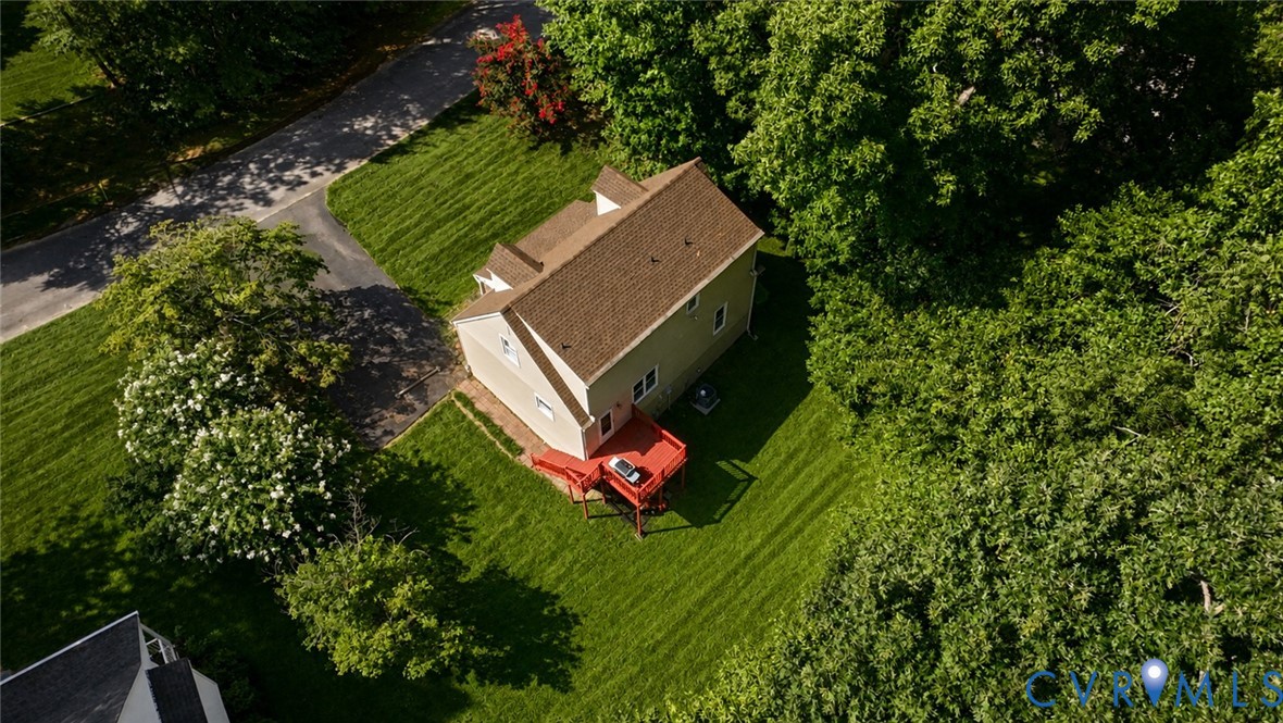 Aerial perspective featuring a multi-story residence with an elevated deck, paved driveway, and expansive lawn surrounded by mature trees