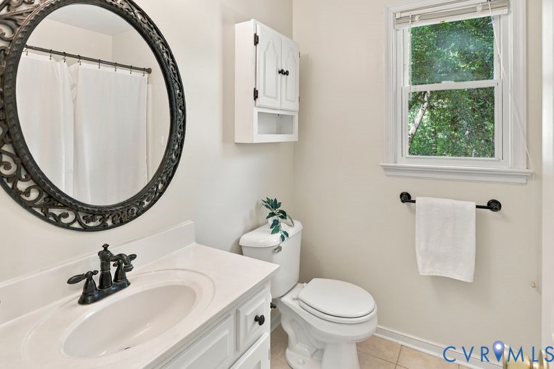 White vanity featuring a smooth-surface countertop, integrated oval basin, and matte black widespread faucet, paired with a decorative circular framed mirror, wall-mounted cabinet, and window with natural light