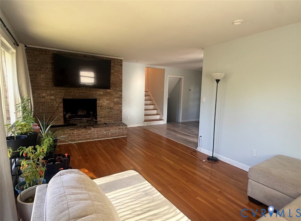 Living area featuring wood-finish flooring, a floor-to-ceiling brick fireplace with a raised brick hearth, and a wooden staircase