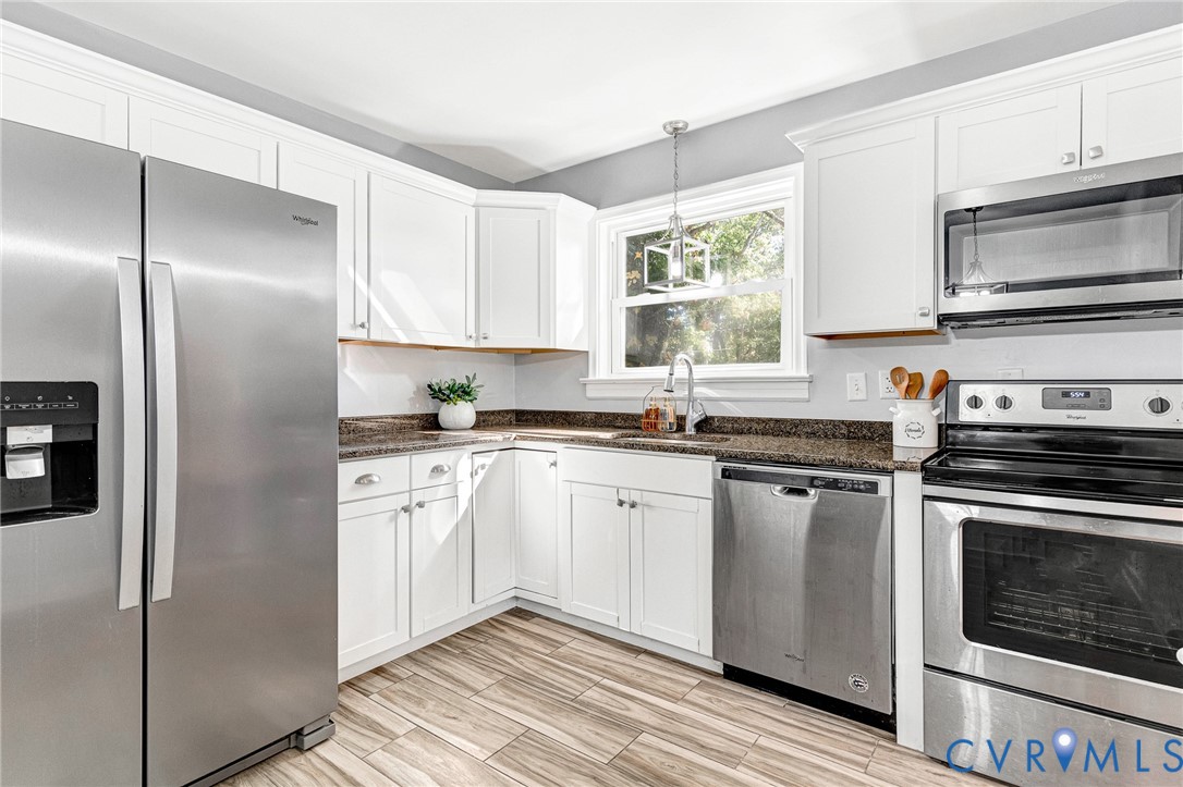 Kitchen featuring white cabinetry, wood-finish flooring, dark stone countertops, stainless steel appliances, and a geometric pendant light fixture Kitchen featuring white cabinetry, wood-finish flooring, dark stone countertops, stainless steel appliances, and a geometric pendant light fixture