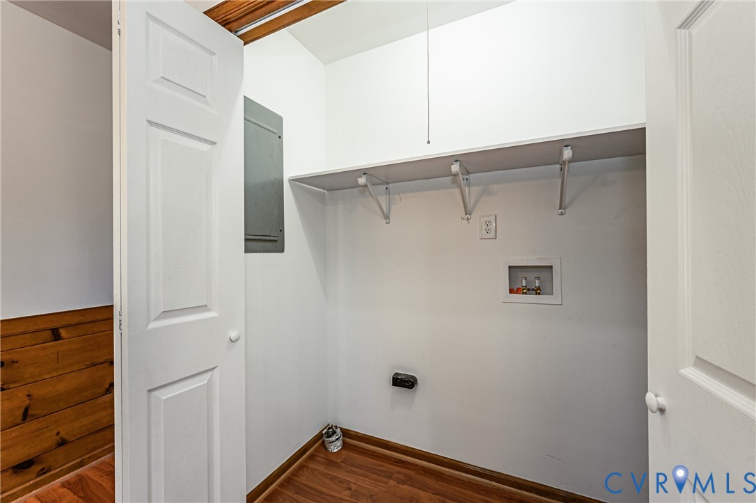 Utility closet featuring wood-finish flooring, a white overhead storage shelf with bracket supports, a recessed plumbing hookup box, and an electrical service panel