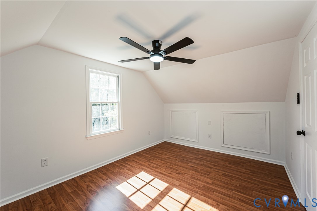 Room featuring sloped ceilings, wood-finish flooring, a multi-blade ceiling fan with an integrated light fixture, and two built-in access panels