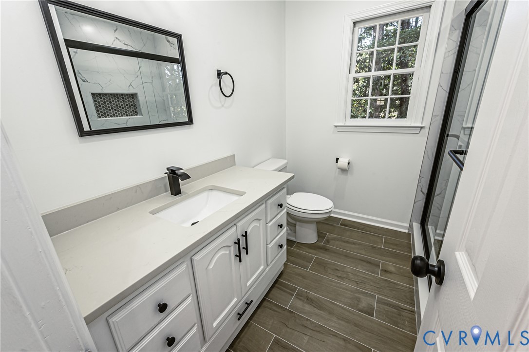 Bathroom featuring a white vanity with a composite countertop and integrated sink, matte black hardware, wood-finish tile flooring, a rectangular mirror with a black frame, and a shower with a glass enclosure