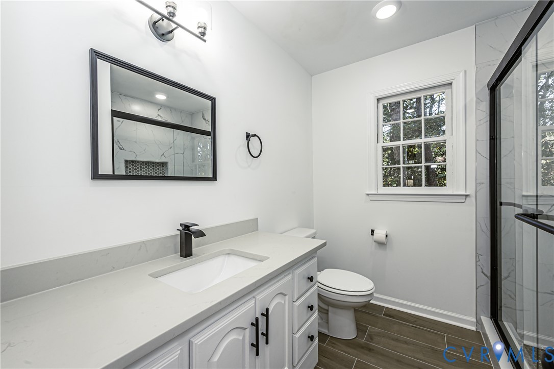 Bathroom featuring a vanity with a light-toned solid surface countertop, rectangular undermount sink, matte black hardware, wood-finish flooring, and a walk-in shower with floor-to-ceiling tile surround