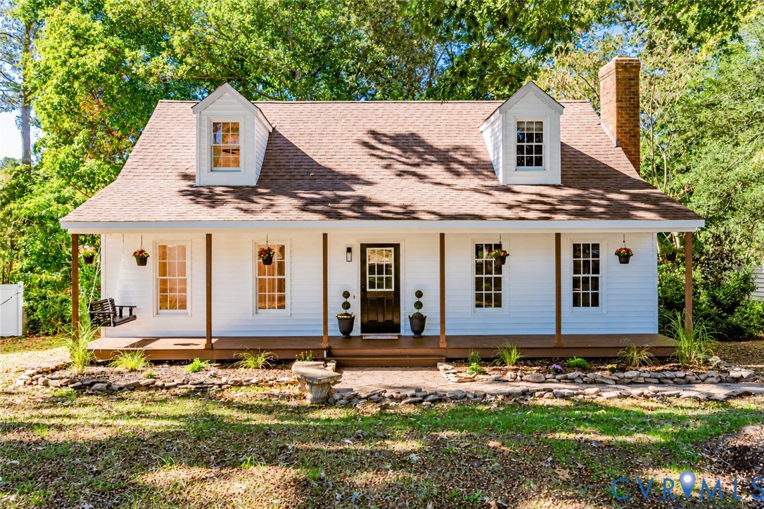 Cape Cod style exterior featuring white siding, a covered front porch with wood decking, dual dormer windows, and a masonry chimney