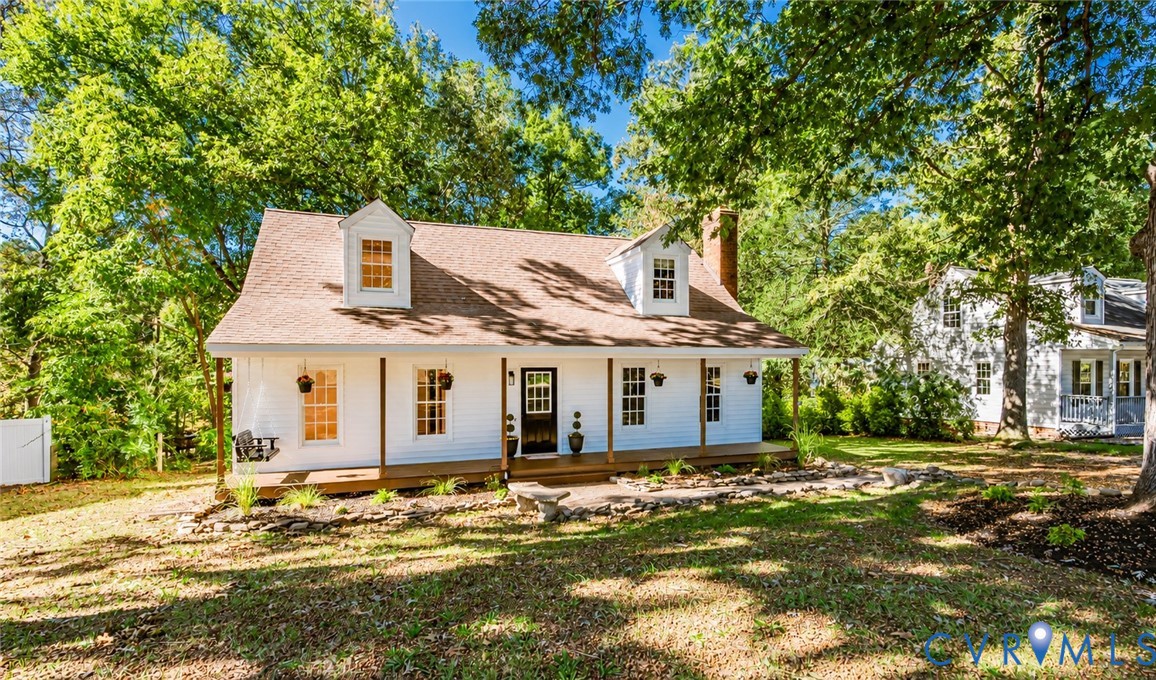 Cape Cod-style residence featuring white siding, a full-length covered front porch, dual dormer windows, a brick chimney, and a stone-lined landscaping bed