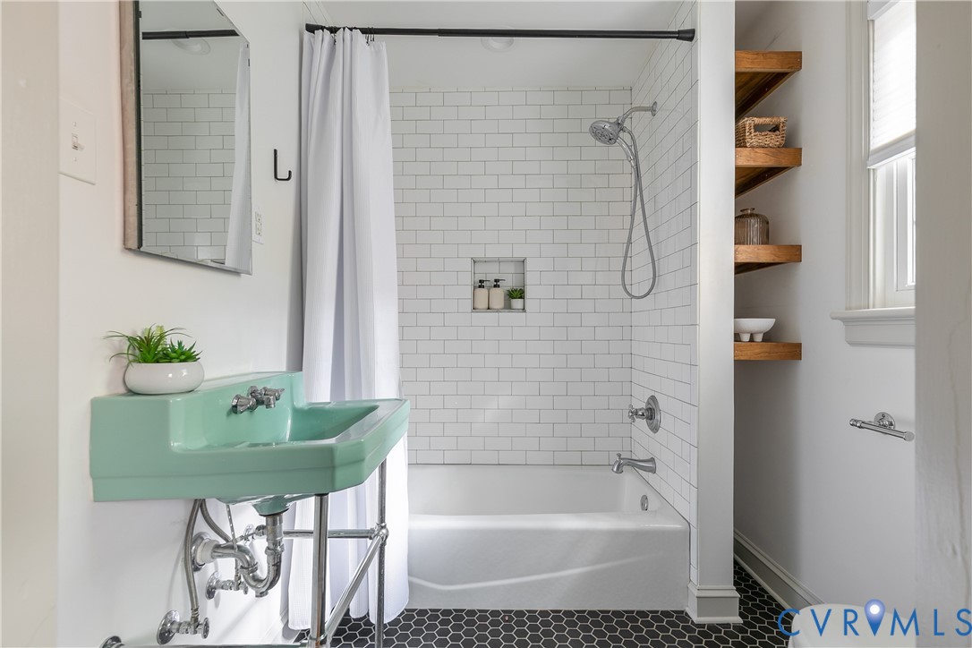 Wall-mounted console sink with exposed plumbing, white subway tile shower surround with integrated soap niche, black hexagonal floor tile, and wood-finish built-in shelving