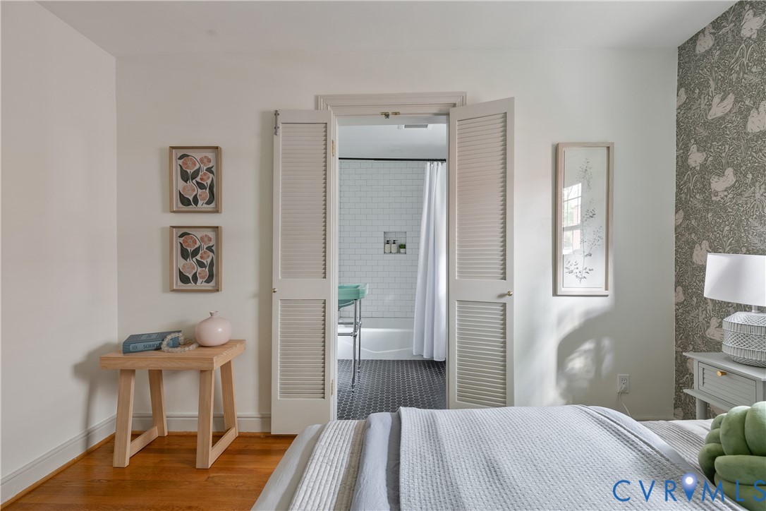 Wood-finish flooring, patterned accent wall covering, and white bi-fold louvered doors opening to a space with tiled flooring and white subway-tiled wall surfaces