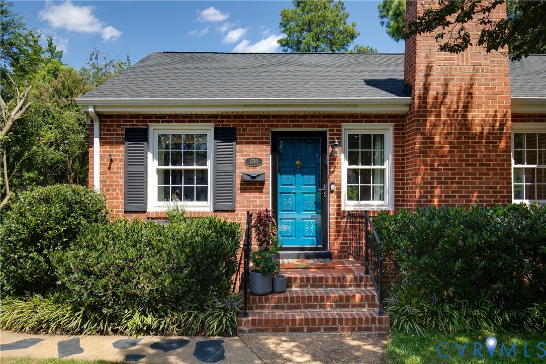 Red brick exterior featuring a blue paneled front door, an external brick chimney, dark shutters, and tiered brick entryway steps with iron handrails