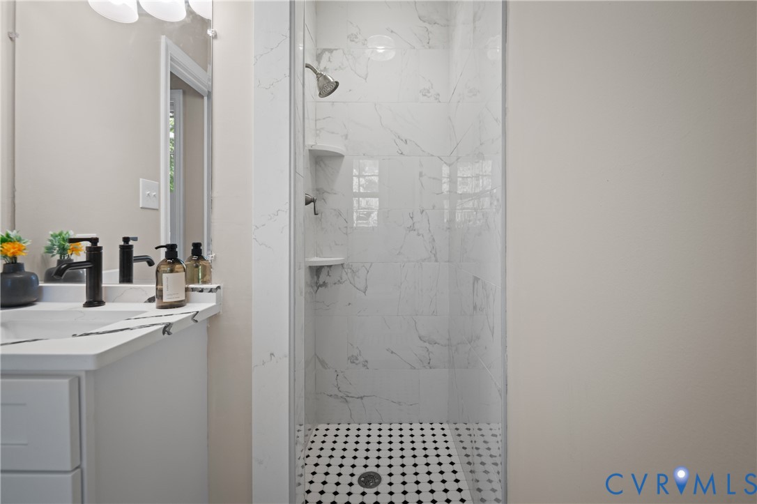 Shower stall featuring large-format veined tile surround and built-in shelving, complemented by a mosaic tile floor, white vanity with stone-finish countertop, and dark-finish faucet fixtures