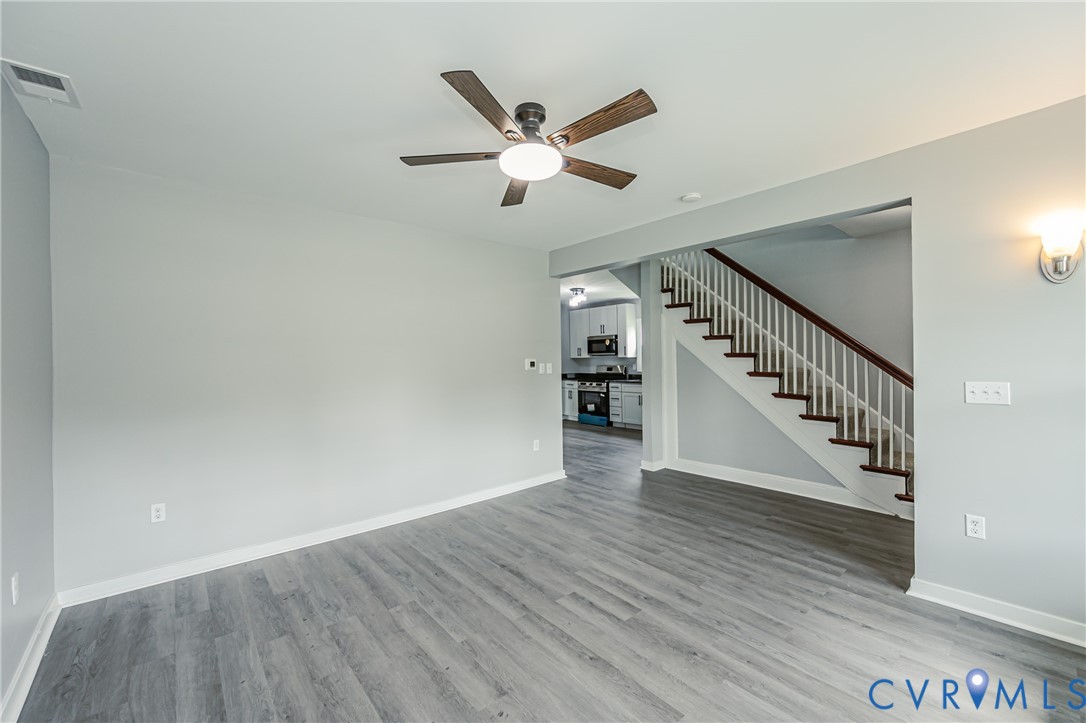 Neutral-toned interior featuring wood-finish flooring, a ceiling fan with integrated lighting, a wall-mounted sconce, and a staircase with a wooden handrail and white balusters