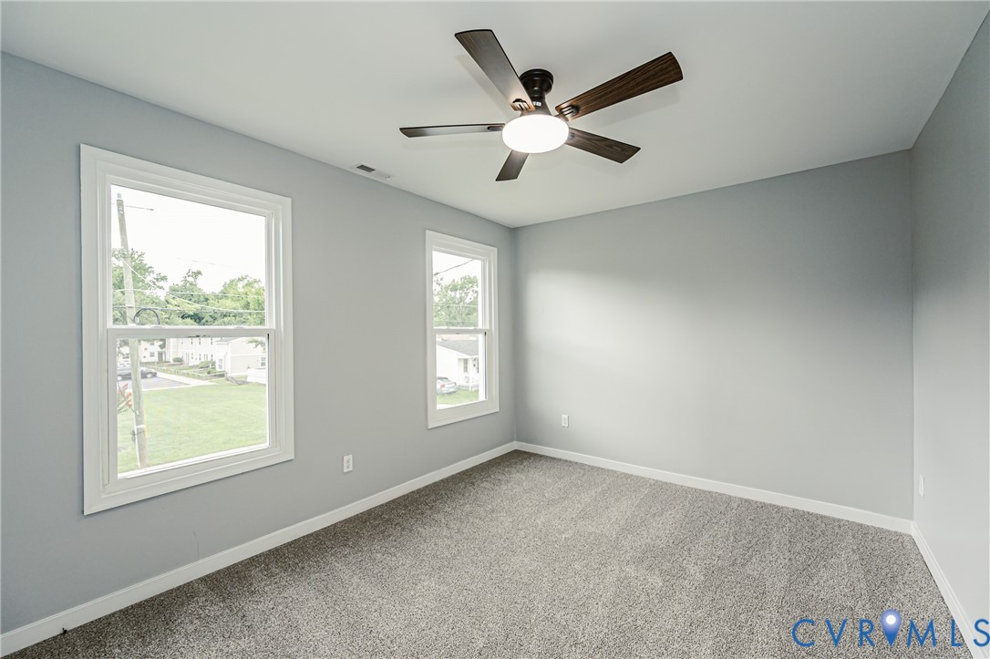 Neutral-toned interior featuring plush carpeting, dual double-hung windows, and a dark wood-finish ceiling fan with integrated lighting