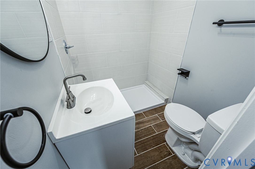 White vanity featuring an integrated sink with a brushed-finish faucet, walk-in shower with horizontal tile surround, wood-finish flooring, and matte black hardware