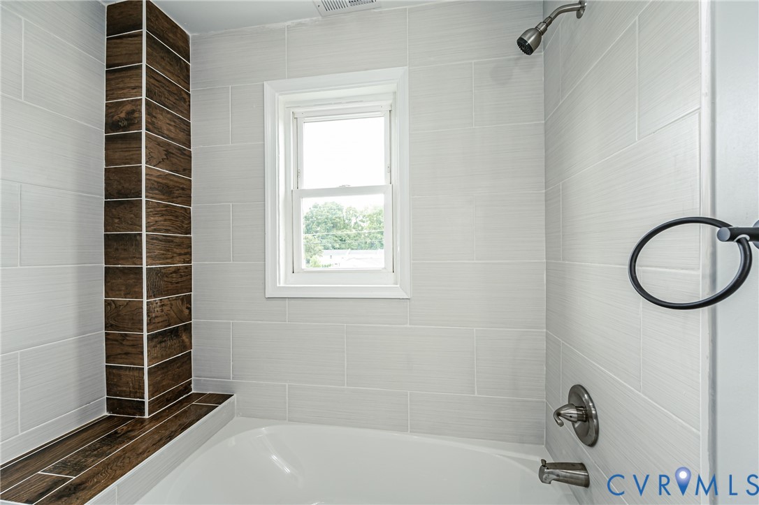Bathtub surround featuring light gray horizontal wall tile, wood-finish accent tiling, integrated ledge, chrome plumbing fixtures, and a single-hung window