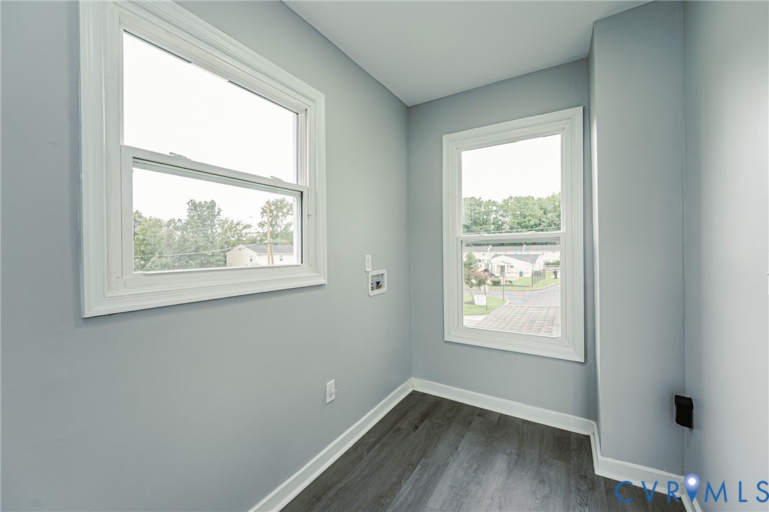 Neutral-toned interior space featuring wood-finish flooring, two double-hung windows, and utility hookups