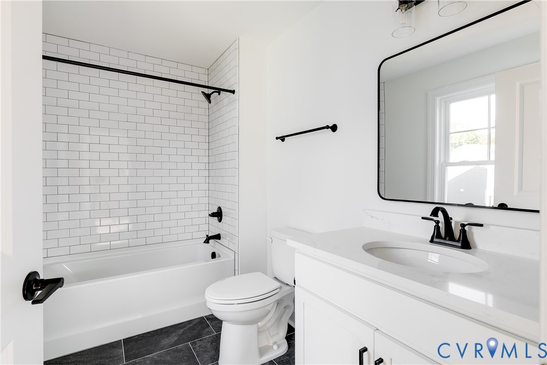 Bathroom featuring a tub-shower combination with white subway tile surround, vanity with a white countertop and oval undermount sink, matte black hardware, and dark tile flooring