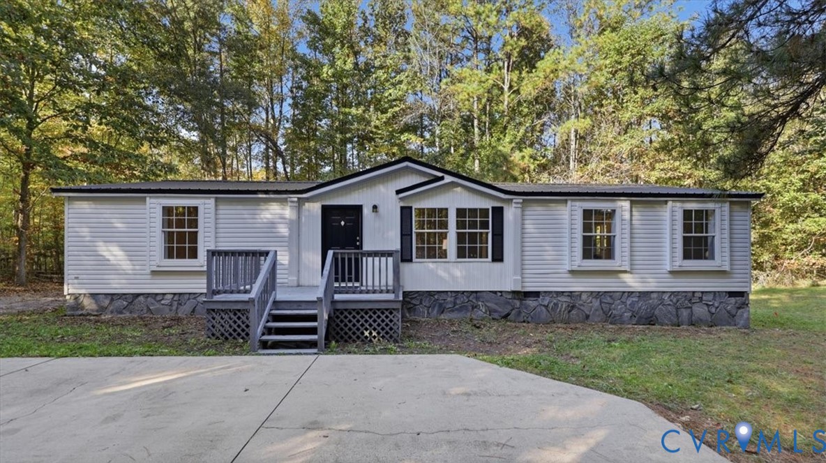 Single-story exterior featuring light-colored horizontal siding, a stone veneer foundation, dark-trimmed window shutters, a front wooden deck with stairs, and a concrete driveway