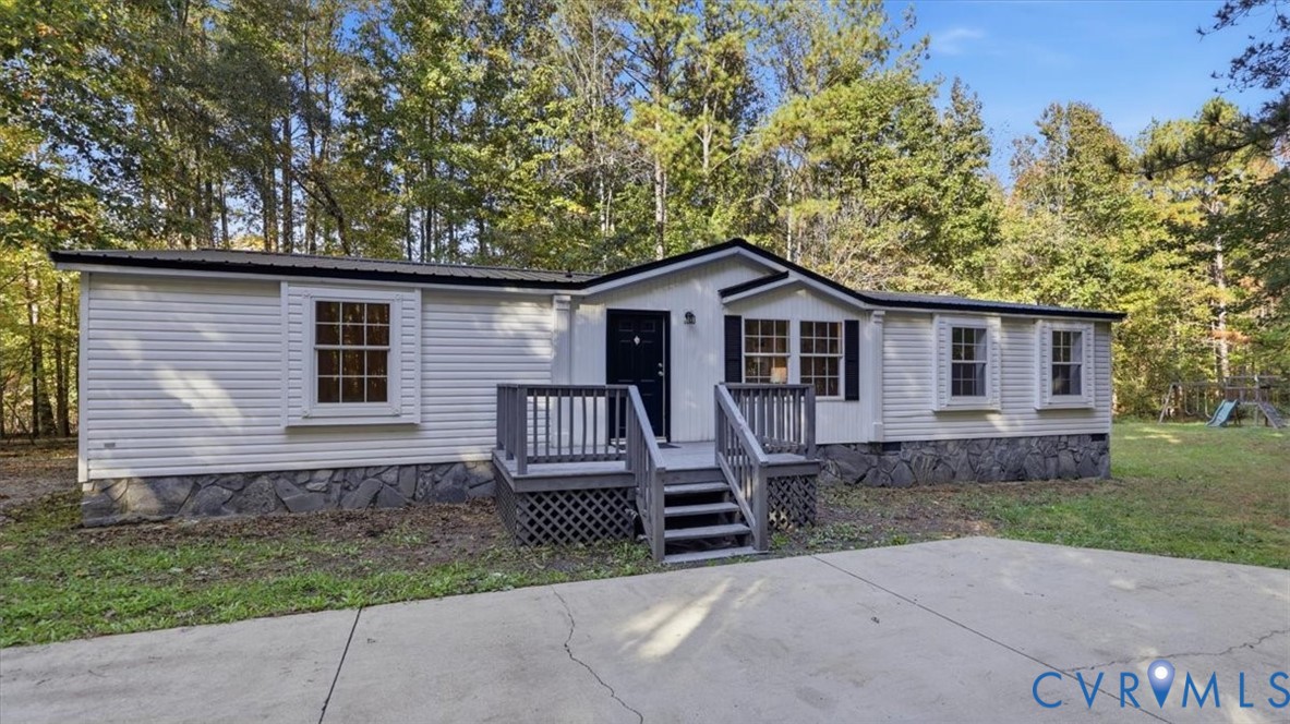 Single-story exterior featuring white horizontal siding, stone-veneer skirting, a wooden deck with lattice foundation, and a concrete driveway Single-story exterior featuring white horizontal siding, stone-veneer skirting, a wooden deck with lattice foundation, and a concrete driveway