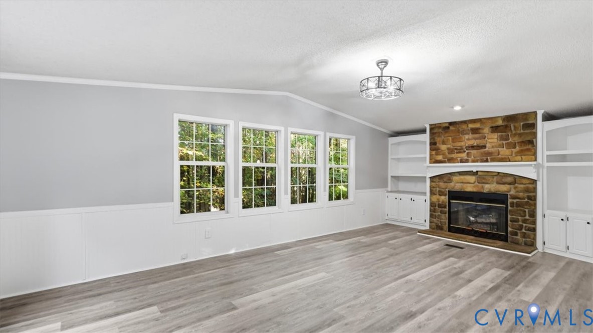Living area featuring stone-clad fireplace, wood-finish flooring, built-in shelving, white wainscoting, and multi-pane windows