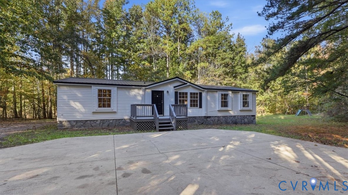 Single-story exterior featuring light-toned horizontal siding, stone-patterned skirting, a metal roof, and a raised wooden front deck