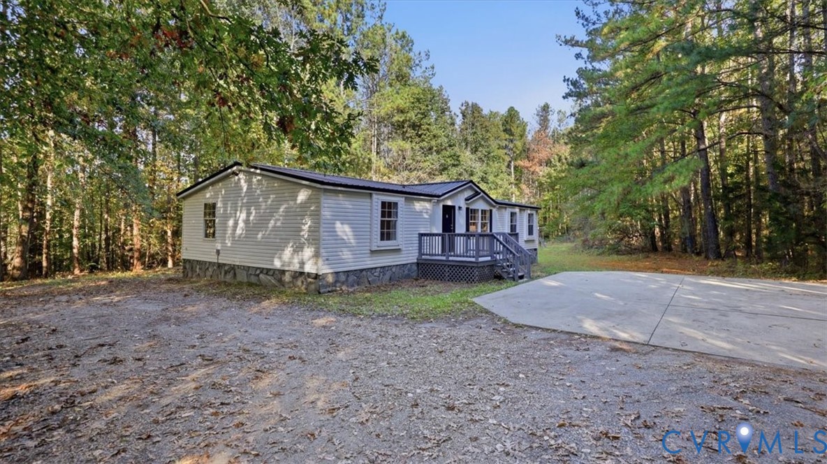 Single-story residence featuring light-colored siding, stone-veneer skirting, a covered deck, and a paved concrete pad, situated on a wooded lot