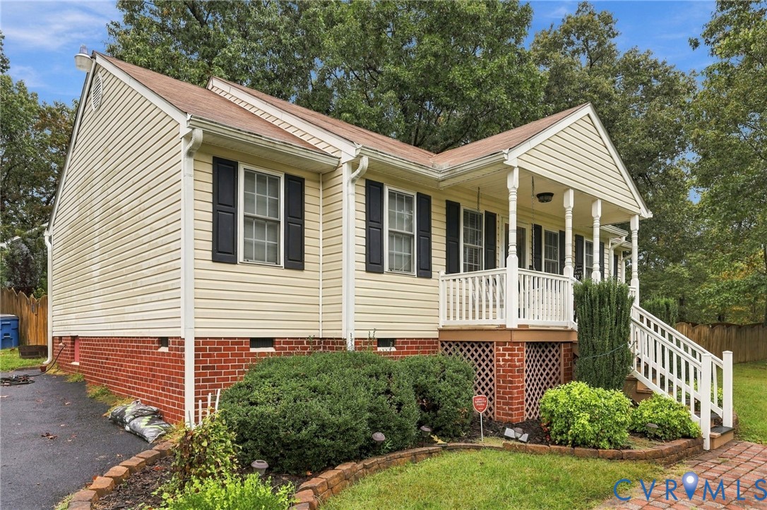 Exterior featuring tan vinyl siding, a brick foundation, a covered front porch with decorative railings, and a landscaped yard with mature trees