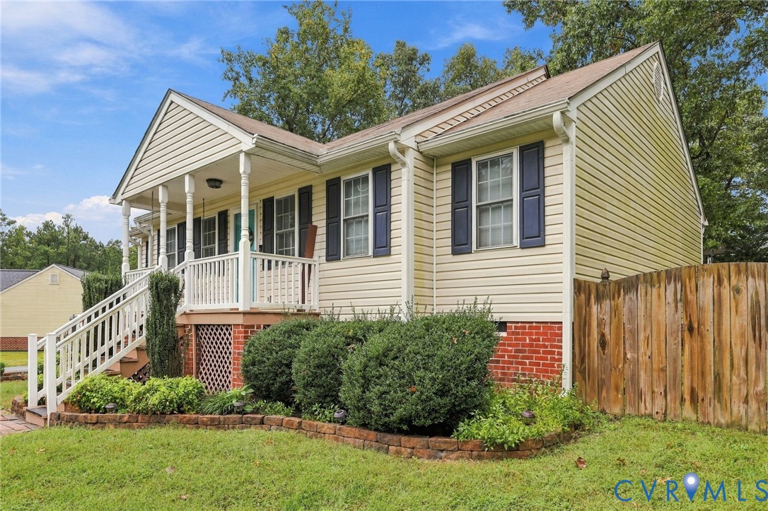 Exterior featuring light-colored vinyl siding, a front porch with white railing, brick foundation detailing, dark shutters, and a privacy fence