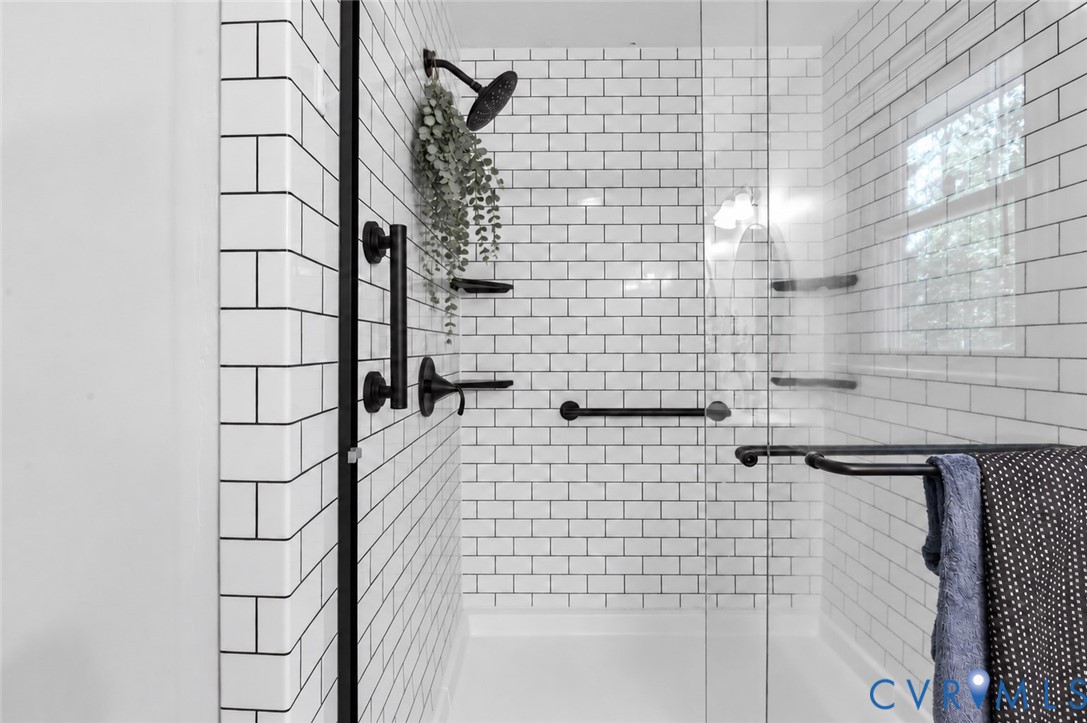 Walk-in shower featuring white subway tile surround, matte black rainfall showerhead, glass door enclosure, and integrated black metal grab bars