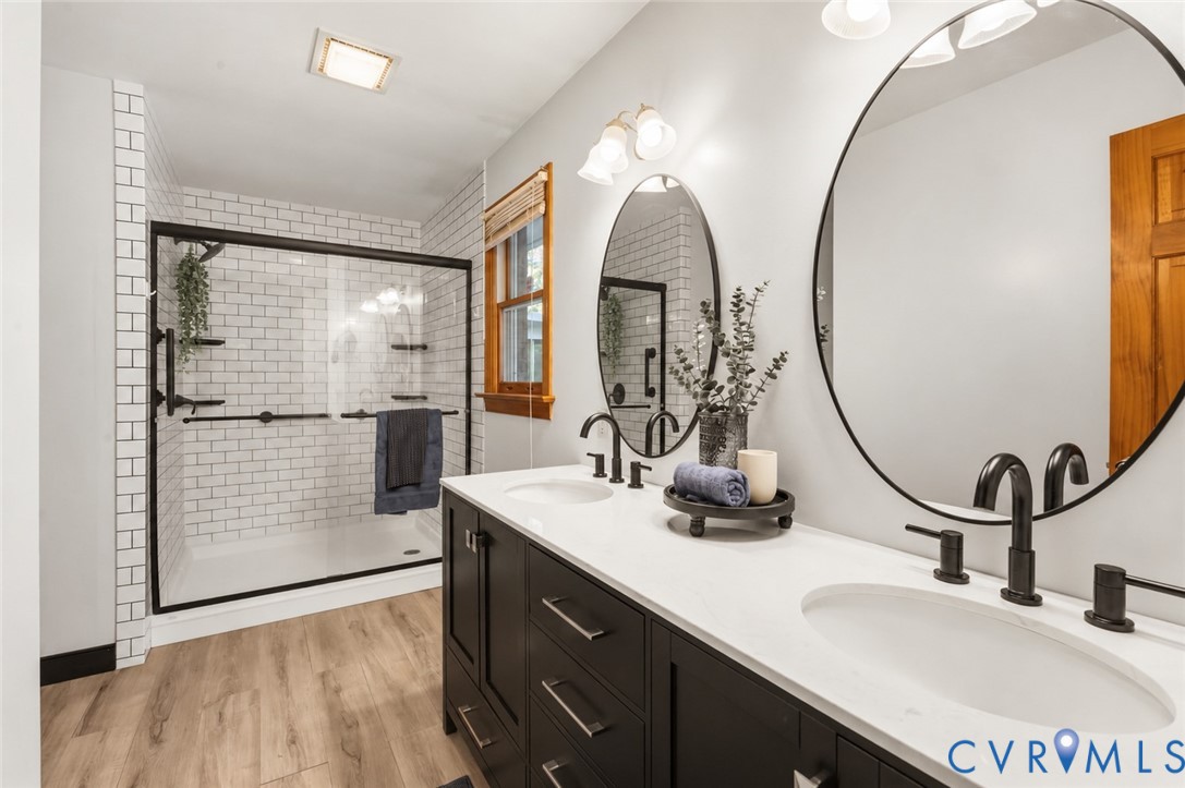 Dual-sink vanity with matte black fixtures, white stone-composite countertop, subway tile shower enclosure with glass door, and wood-finish flooring