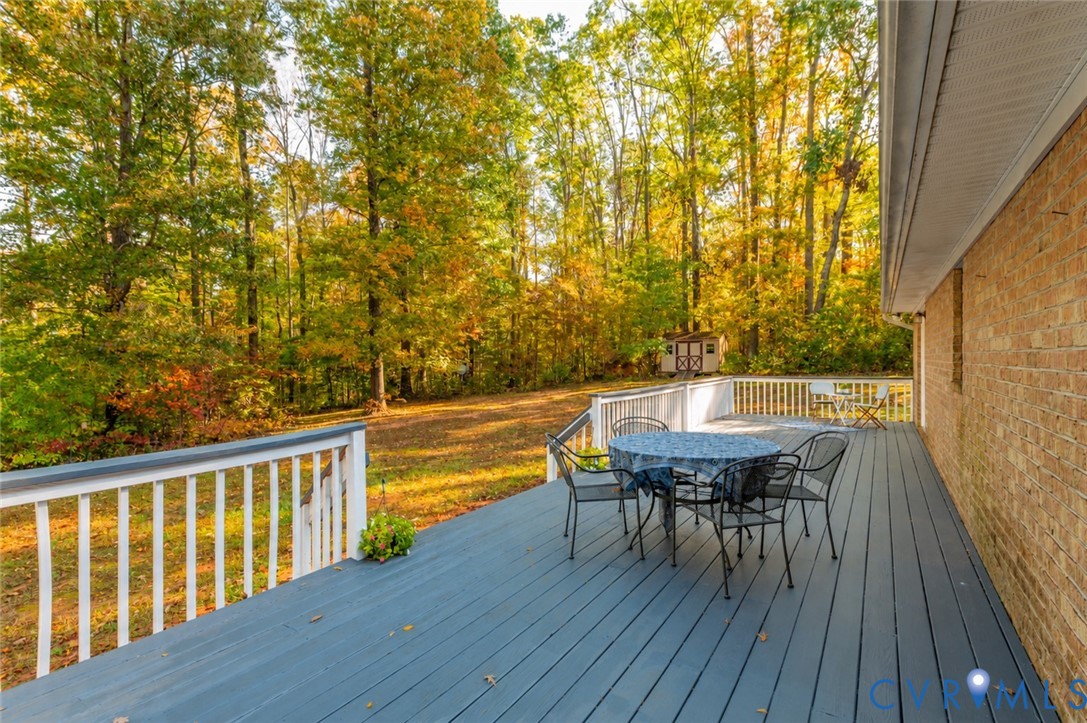 Expansive deck featuring painted wood-finish planks, white railings, and a brick exterior wall, set against a backdrop of mature trees
