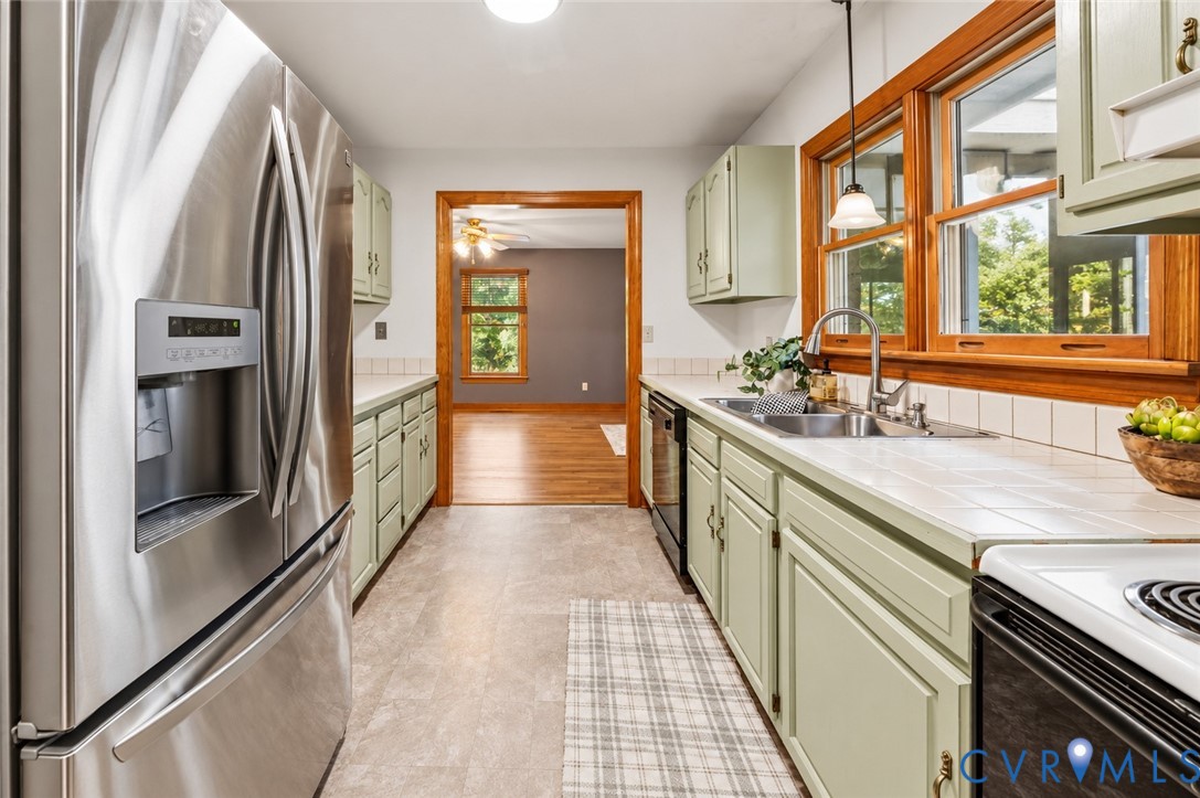 Kitchen featuring stainless steel refrigerator, tile countertops, wood-finished cabinetry, double-basin sink with gooseneck faucet, and wooden window trim