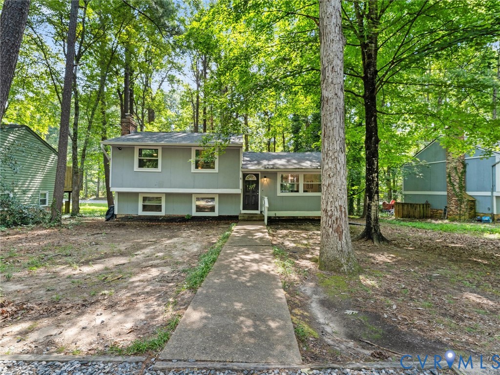 Split-level exterior featuring light gray horizontal siding, a central front entry, concrete walkway, and mature wooded surroundings