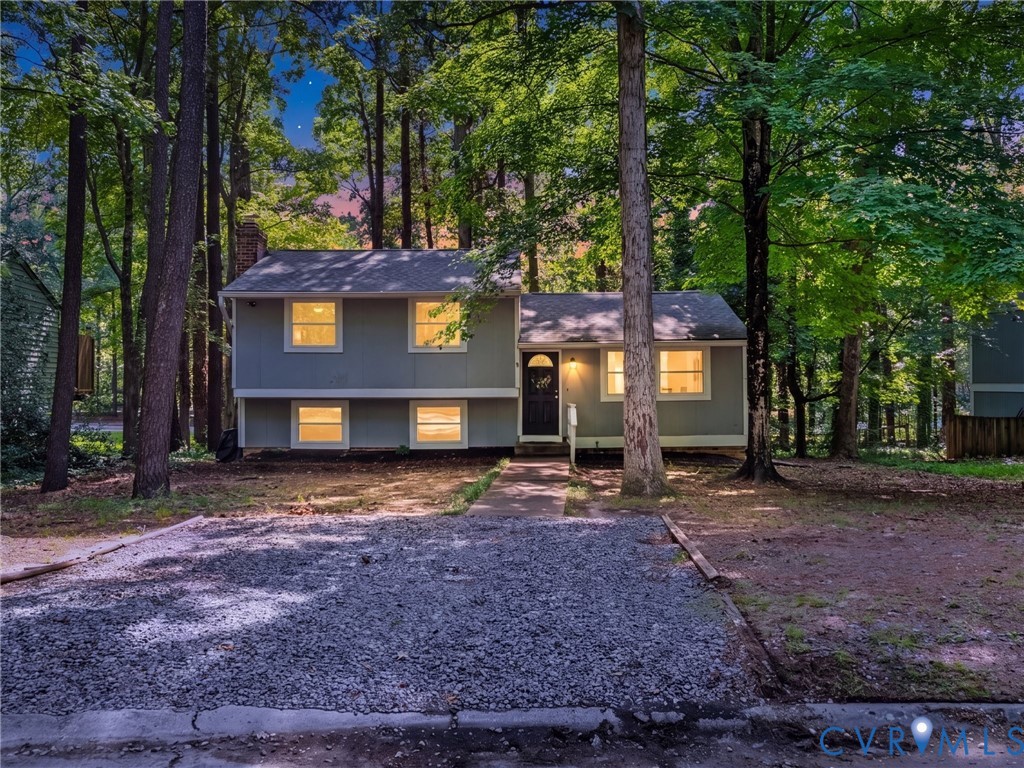 Split-level exterior featuring grey siding, a front-facing brick chimney, concrete walkway, gravel parking area, and mature perimeter trees
