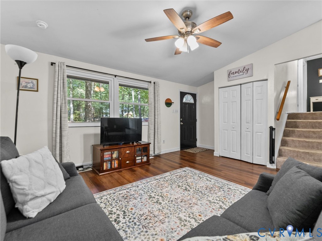 Living area featuring wood-finish flooring, a multi-blade ceiling fan with integrated light fixture, dual-panel closet doors, and windows with multi-pane glass