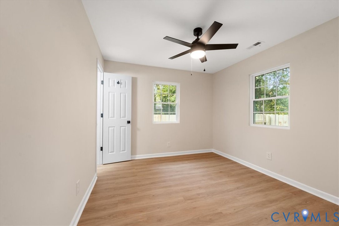 Neutral-toned interior space featuring light wood-finish flooring, dual-pane windows, a six-panel door, and a matte black ceiling fan with an integrated light fixture