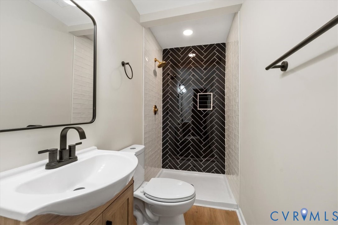 Bathroom featuring a walk-in shower with black herringbone-patterned tile, wood-finish vanity with white basin, matte black hardware, and an arched-frame mirror