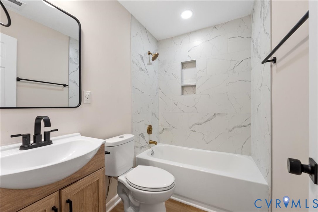 Bathroom featuring a wood-finish vanity with a white vessel sink, matte black faucet, wall-mounted mirror, and a tub-shower combination with floor-to-ceiling surround and built-in recessed shelving
