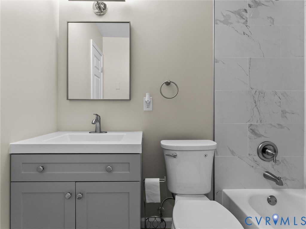 Grey vanity with a white countertop and rectangular sink, square mirror, chrome faucet, and tile surround bathtub