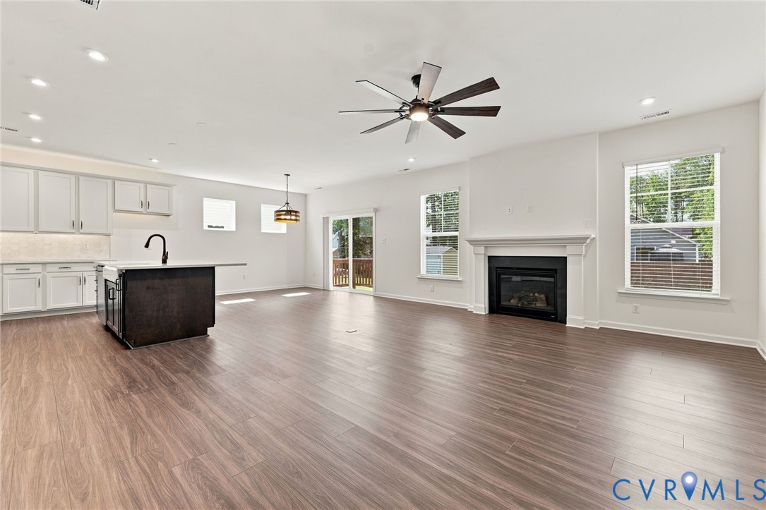 Open-concept interior featuring wood-finish flooring, a kitchen island with dark cabinetry, recessed lighting, a fireplace with a white mantel, and a ceiling fan