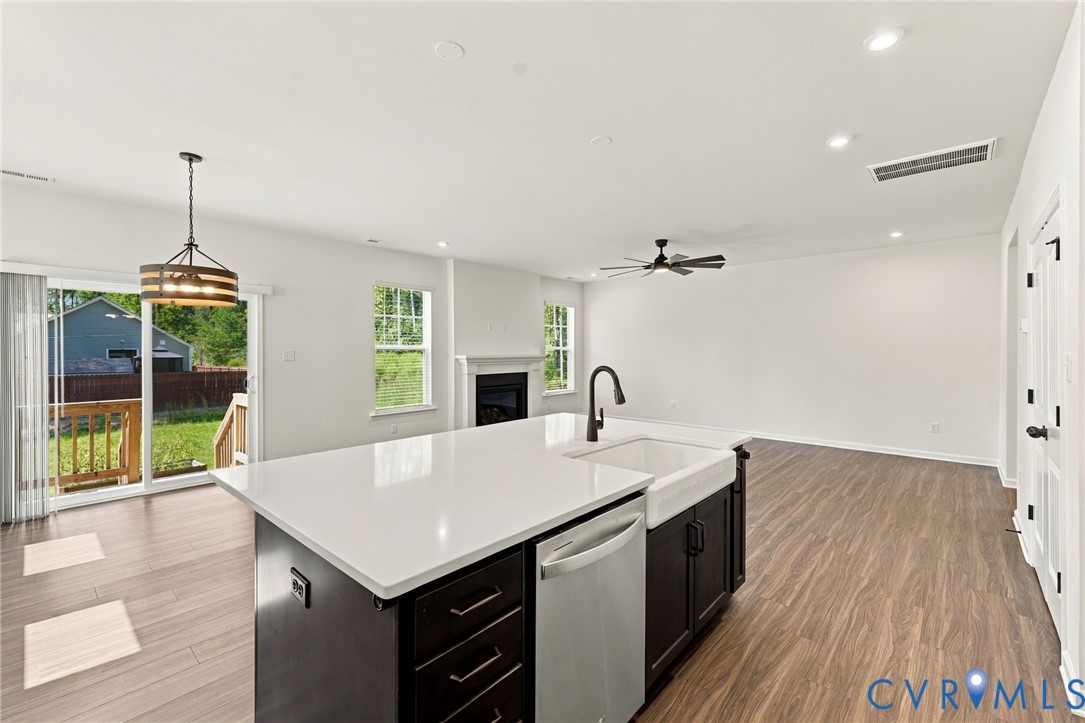 Kitchen island featuring a white surface, dark cabinetry, and an apron-front sink, wood-finish flooring, recessed lighting, and a fireplace with a decorative mantel