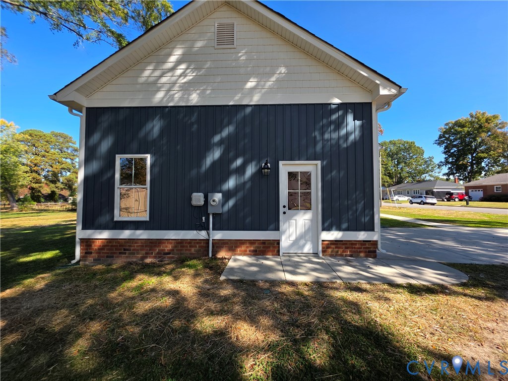 Exterior featuring vertical board-and-batten siding, shake shingle gable, brick base, concrete pad, and white-trim door