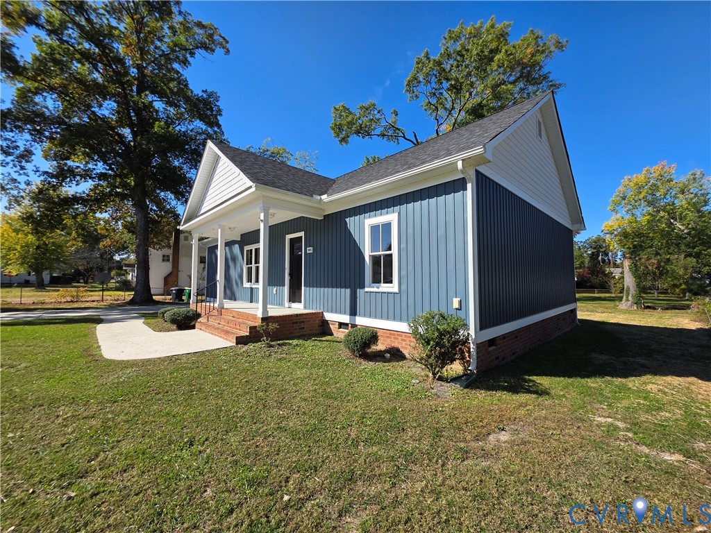 Single-story residence featuring vertical siding, a brick foundation, a covered front porch, and a concrete walkway Single-story residence featuring vertical siding, a brick foundation, a covered front porch, and a concrete walkway